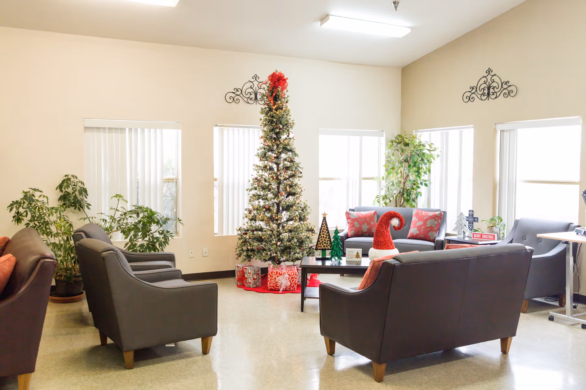 A bright and cozy living room decorated for Christmas with a tall Christmas tree adorned with lights and a red bow on top. The room has several gray armchairs and sofas with red and floral cushions arranged around a black coffee table. There are potted plants near the windows, and holiday decorations including a Santa hat and small Christmas trees on the coffee table. The walls are light-colored with decorative metal wall art above the windows.