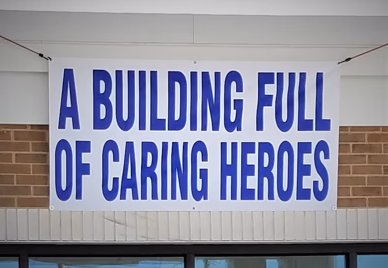 A large white banner with bold blue text that reads 'A BUILDING FULL OF CARING HEROES' hanging on a brick and white wall above a window.