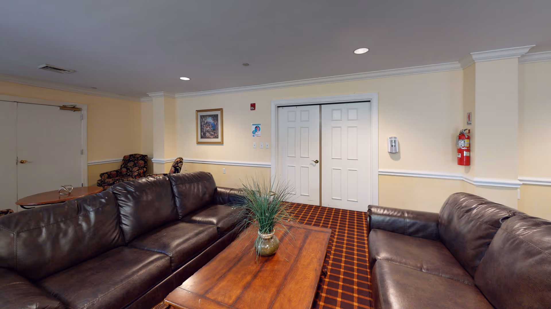 A cozy living room area with two dark brown leather sofas facing each other across a wooden coffee table with a green plant in a vase. The room has beige walls, a patterned carpet, a framed painting on the wall, and a fire extinguisher mounted on the right wall near white double doors.