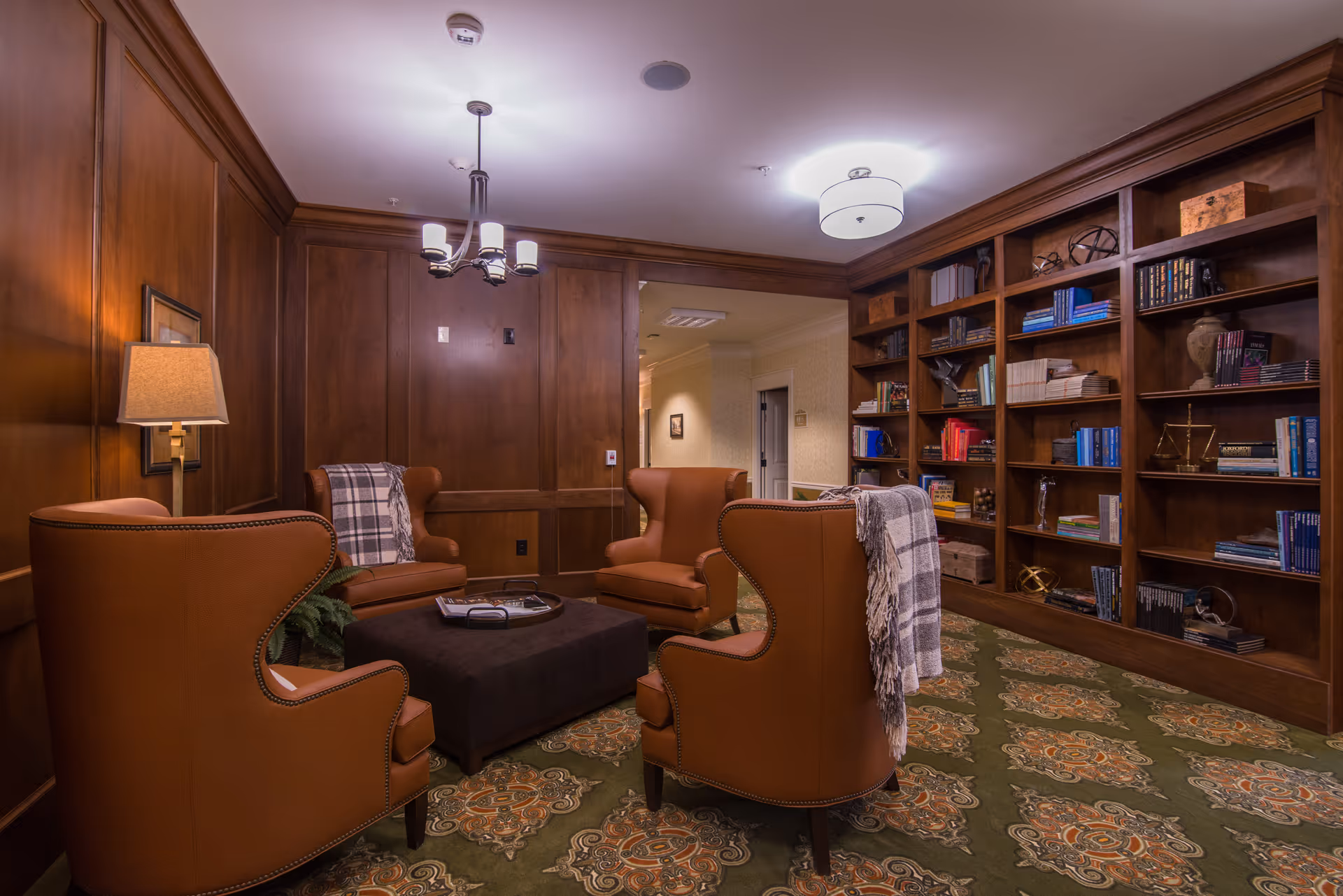 A cozy interior lounge area with four brown leather wingback chairs arranged around a dark ottoman. The room features wood-paneled walls, a patterned carpet, a built-in wooden bookshelf filled with books and decorative items, and soft lighting from a ceiling fixture and a wall lamp.