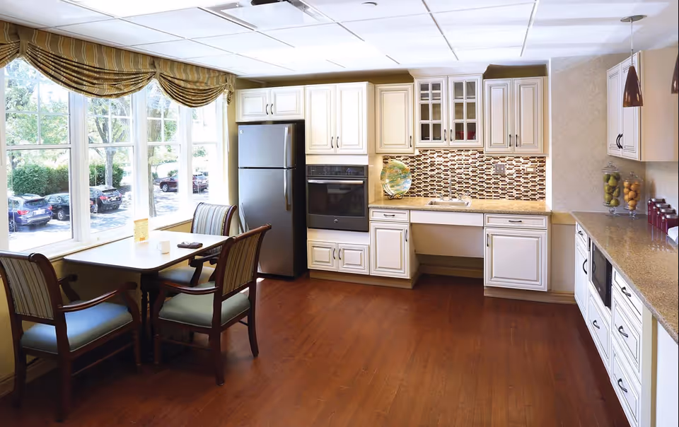 A bright kitchen area with white cabinets, a stainless steel refrigerator, built-in oven, and a mosaic tile backsplash. There is a small dining table with four chairs near large windows with yellow striped valances, allowing natural light to fill the room. The floor is wooden, and the countertop has decorative jars and a plate.