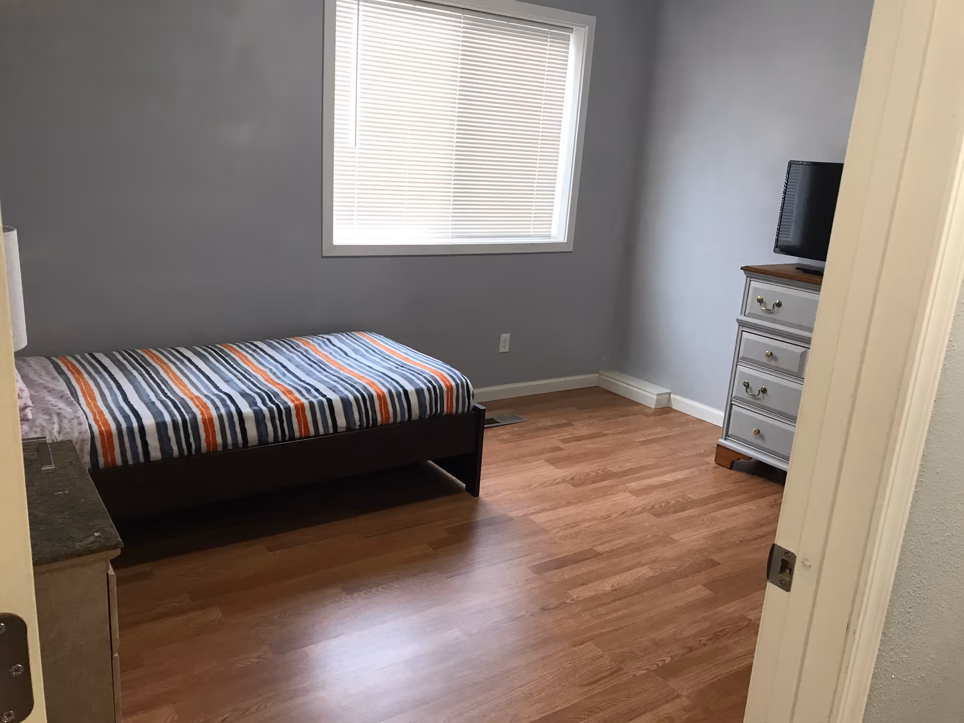 A simple bedroom with a single bed covered in a striped blanket in shades of blue, white, and orange. There is a window with closed blinds on the wall behind the bed. A small dresser with a television on top is positioned against the adjacent wall. The room has light-colored walls and wooden flooring.