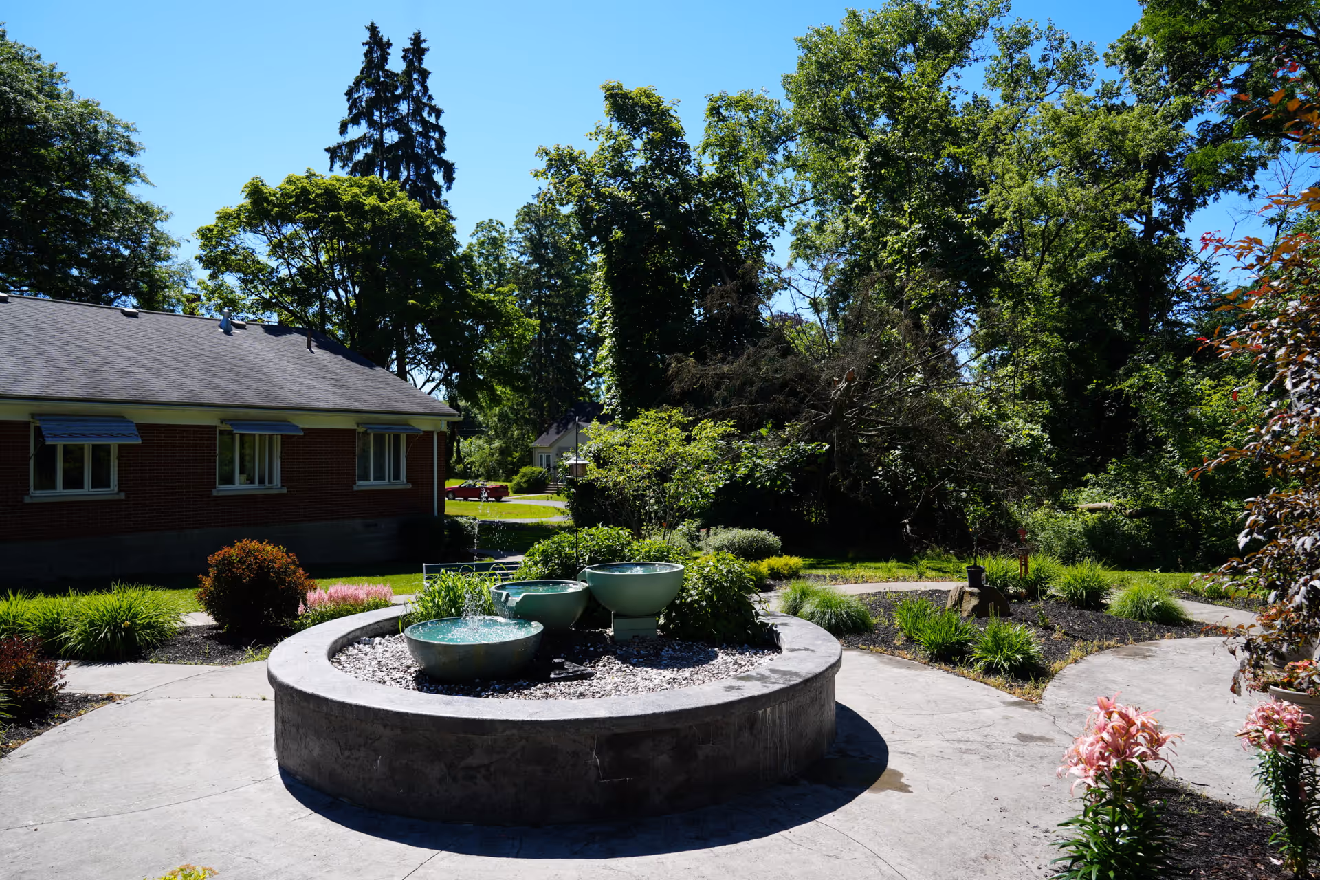 Outdoor garden area with a circular concrete planter featuring three green water bowls with water fountains. Surrounding the planter are pathways, various plants, bushes, and trees. A brick building with windows and awnings is visible on the left side under a clear blue sky.