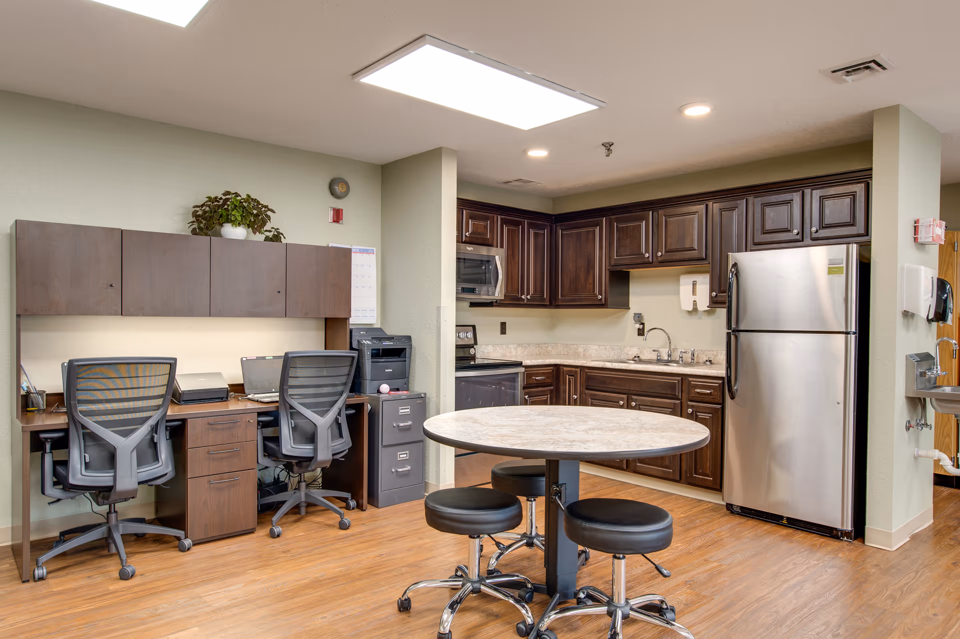 Interior view of a facility room featuring a small kitchen area with dark wood cabinets, a stainless steel refrigerator, a microwave, and a sink. In front of the kitchen is a round table with four black swivel stools. To the left, there is a workspace with two office chairs, a desk, a printer, and overhead cabinets. The floor is wood, and the walls are light-colored.