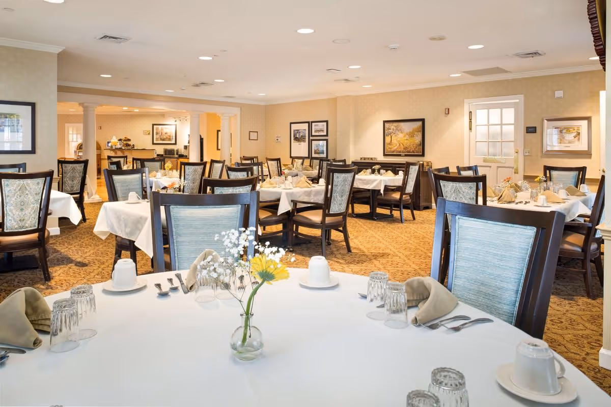 A well-lit dining room in a senior living facility with multiple tables covered in white tablecloths, each set with glasses, cups, napkins, and silverware. The chairs have wooden frames with light blue upholstered backs. The room features beige walls adorned with framed artwork and a patterned carpet. In the background, there is a doorway and a small buffet area.