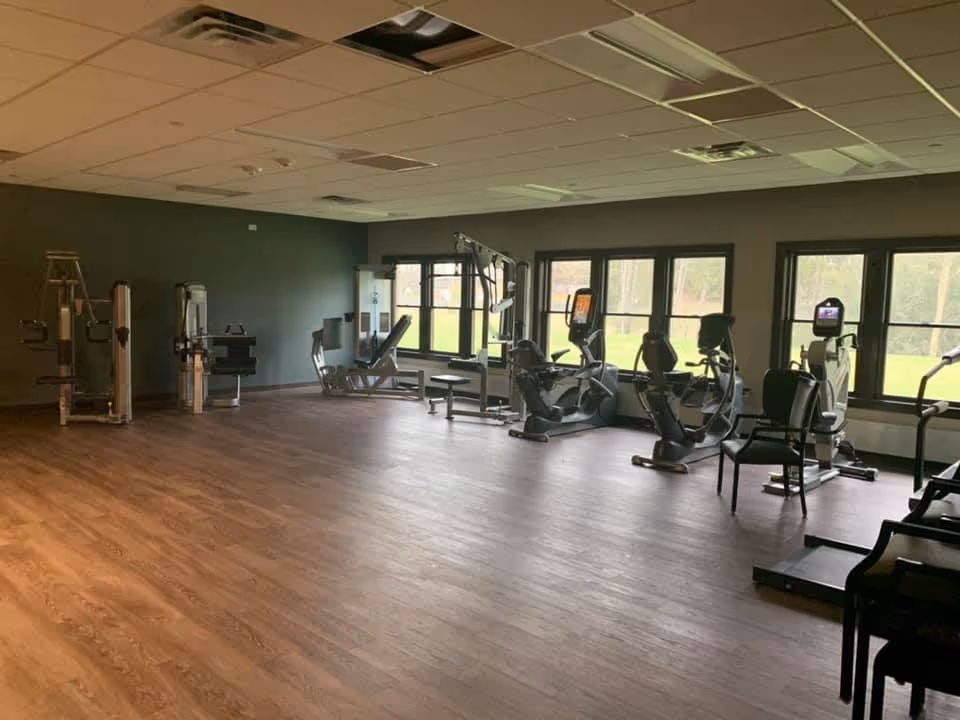 A spacious fitness room with wooden flooring and several exercise machines including stationary bikes, weight machines, and treadmills. Large windows on one side let in natural light and offer a view of greenery outside. The ceiling has recessed lighting and ventilation panels.