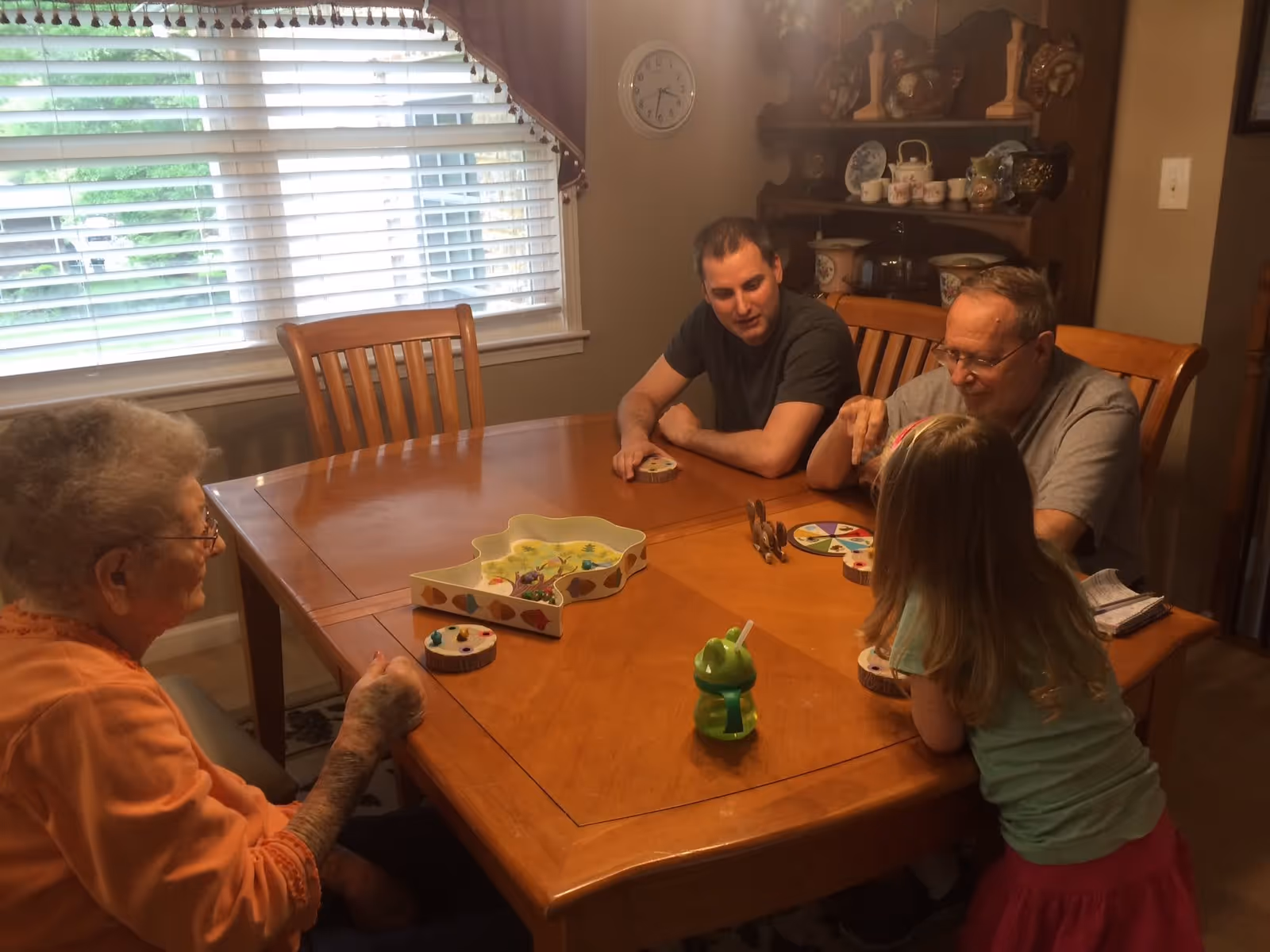 Four people, including an elderly woman, two men, and a young girl, sitting around a wooden dining table playing a board game in a room with a window covered by blinds and a wooden cabinet with decorative items in the background.