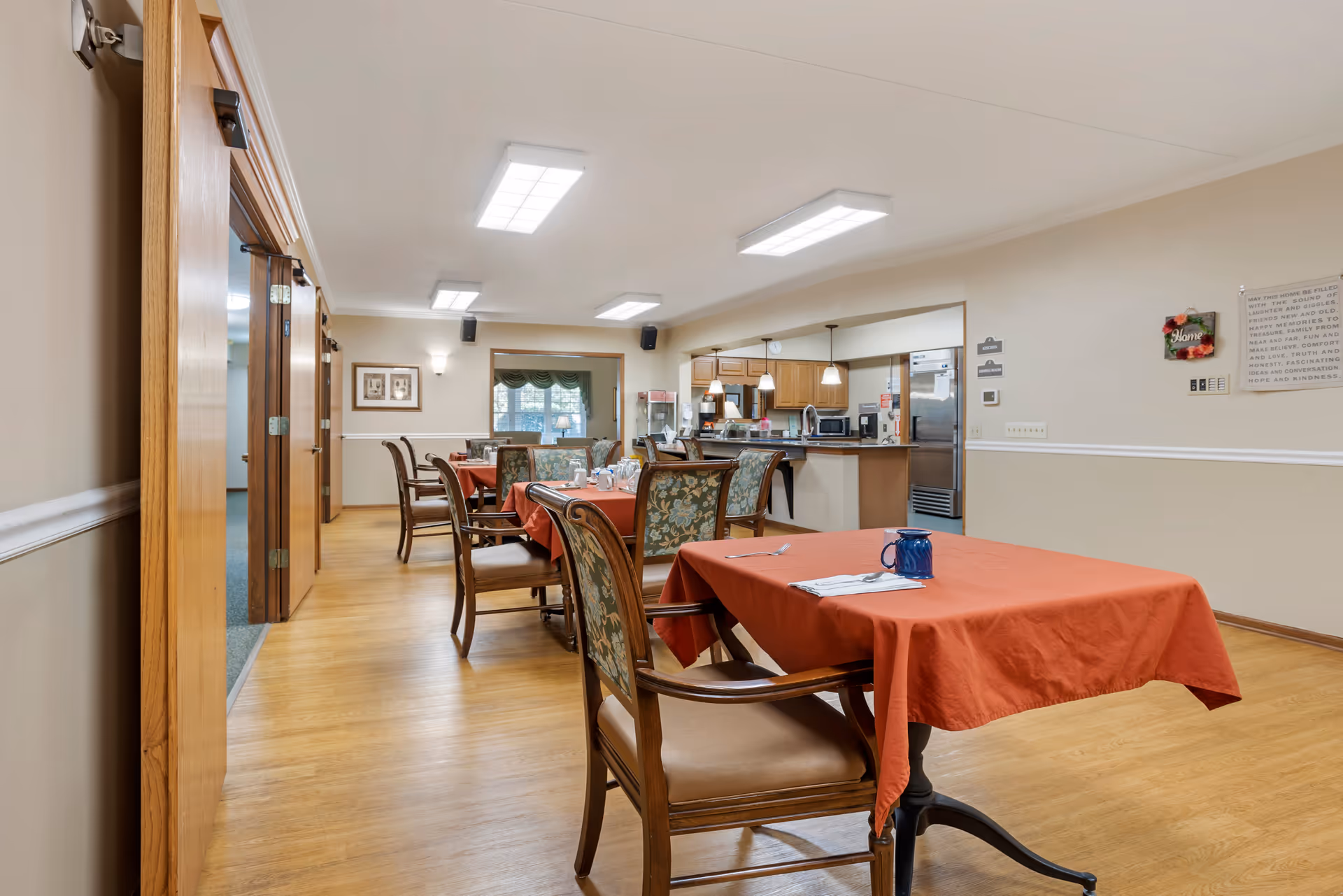 Interior view of a dining area in a senior living facility with wooden floors, tables covered with red tablecloths, and chairs with floral upholstery. The room is well-lit with ceiling lights and has an open kitchen area in the background. There are framed pictures on the wall and a window with green valance curtains.