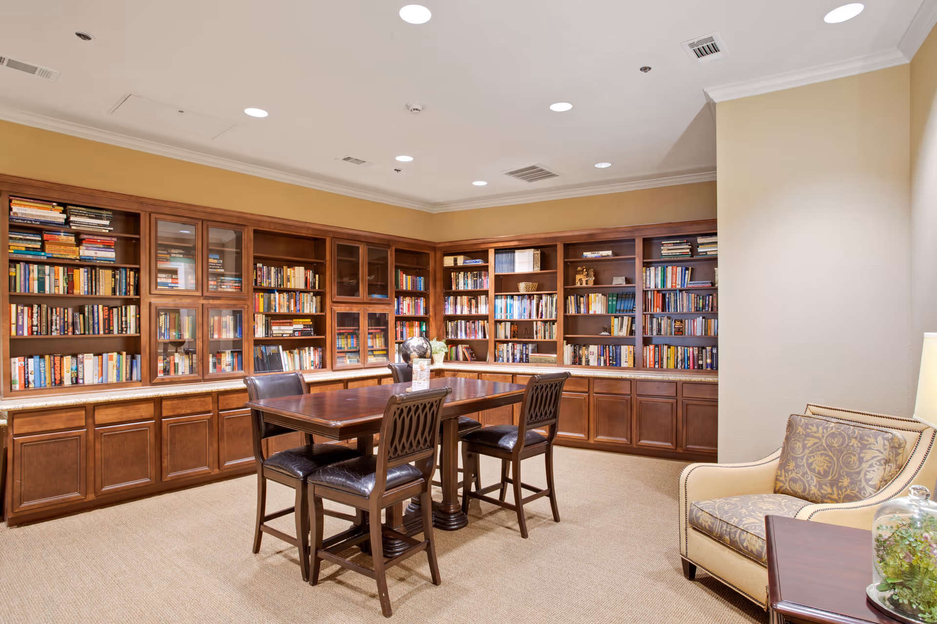 A cozy library room with wooden bookshelves filled with books along two walls. In the center, there is a wooden table with four dark wooden chairs. To the right, there is a beige armchair with a patterned cushion next to a small wooden side table with a glass terrarium on it. The room has beige walls and carpet, and recessed ceiling lights provide illumination.