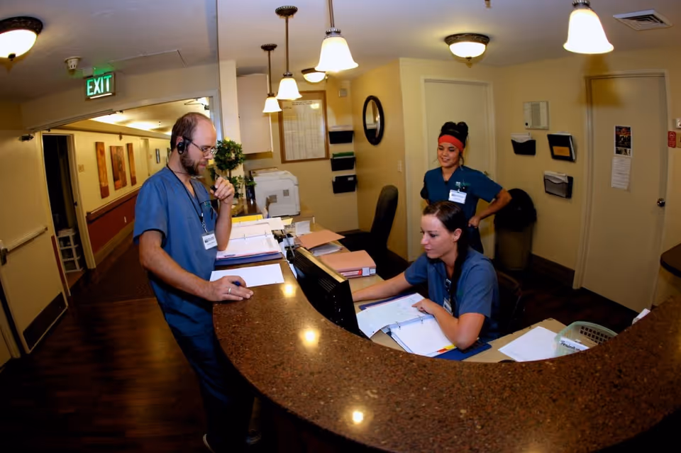 Three healthcare workers in blue scrubs at a reception desk inside a healthcare facility. One man is standing and talking on a headset while looking at papers on the counter. A woman is seated at the desk working on a computer and looking at documents, and another woman stands behind her smiling. The area is well-lit with hanging lights and has a hallway leading to other rooms.