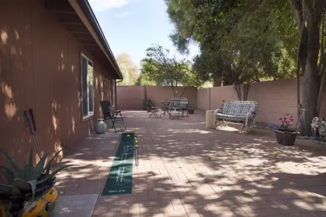 Brick-paved outdoor courtyard with chairs, benches, potted plants and a long practice mat beside a single-story building and a privacy wall.