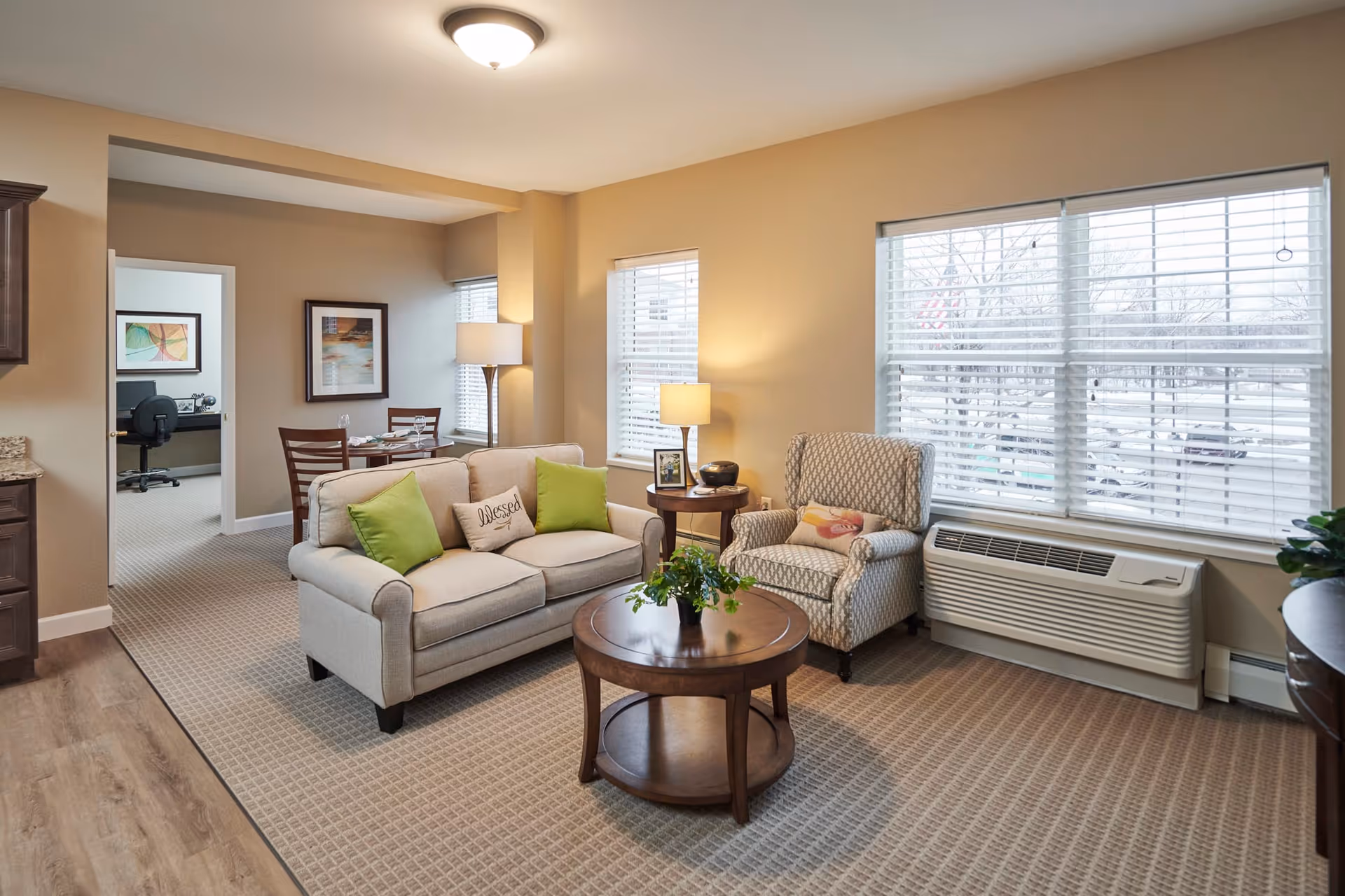 A cozy living room area in a senior living facility with a beige sofa adorned with green and beige pillows, a patterned armchair, a round wooden coffee table with a plant, and two windows with white blinds letting in natural light. A small side table with a lamp and picture frame is next to the armchair. In the background, there is a dining area with a table and chairs, and an office space with a desk and chair is visible through an open doorway.