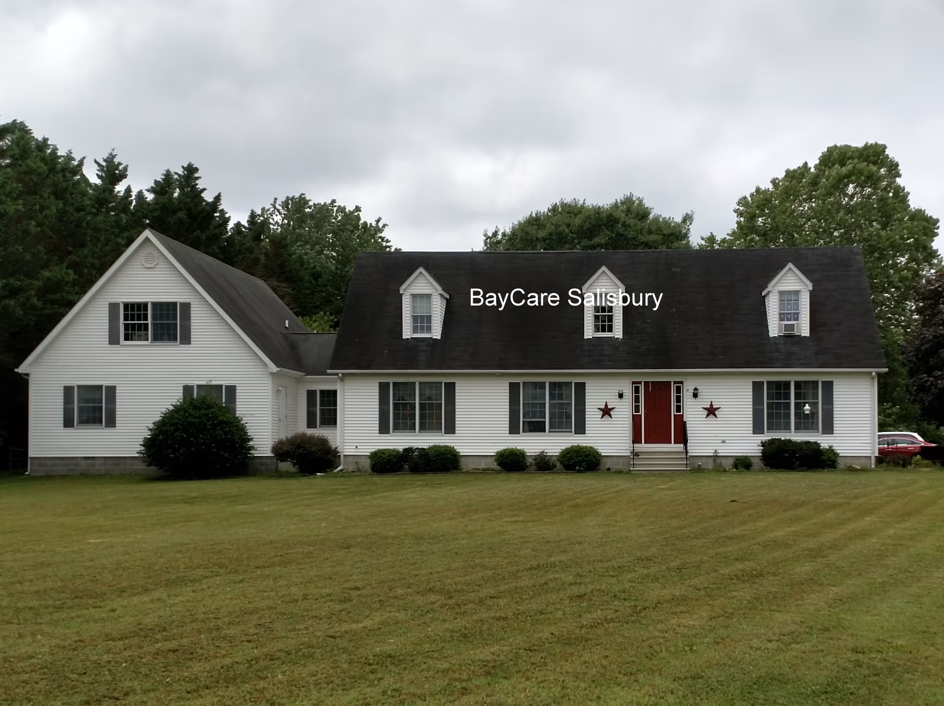 Front view of a white, single-story BayCare assisted living building with a dark roof, red doors, and a well-kept green lawn.