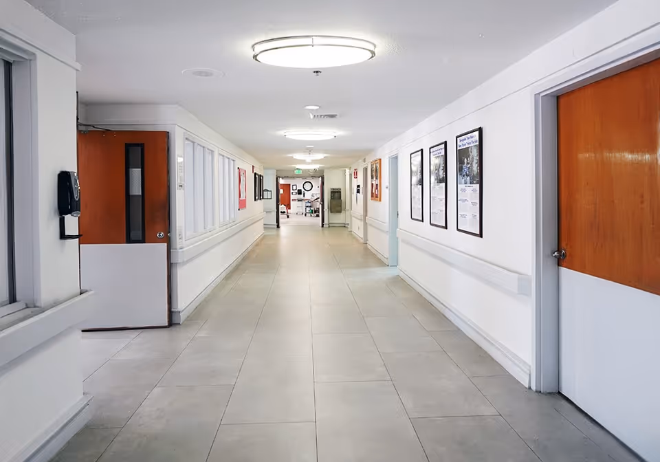 A clean, well-lit hallway in a senior living facility with white walls, tiled floors, and wooden doors on either side. The hallway features handrails along the walls and framed posters or notices. Ceiling lights illuminate the corridor, and there is a water fountain and some equipment visible at the far end.