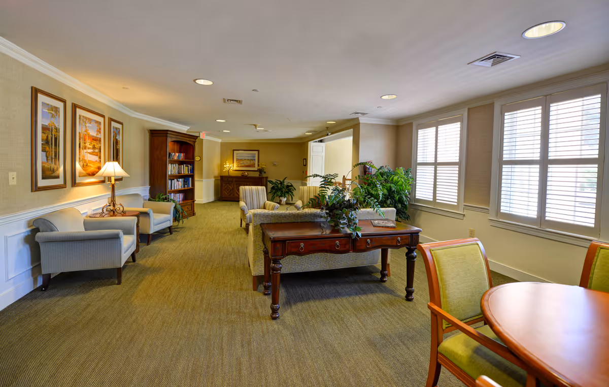 A well-lit common area in a senior living facility featuring comfortable armchairs, a wooden table with plants, framed artwork on the walls, a bookshelf, and large windows with white blinds allowing natural light to fill the room.