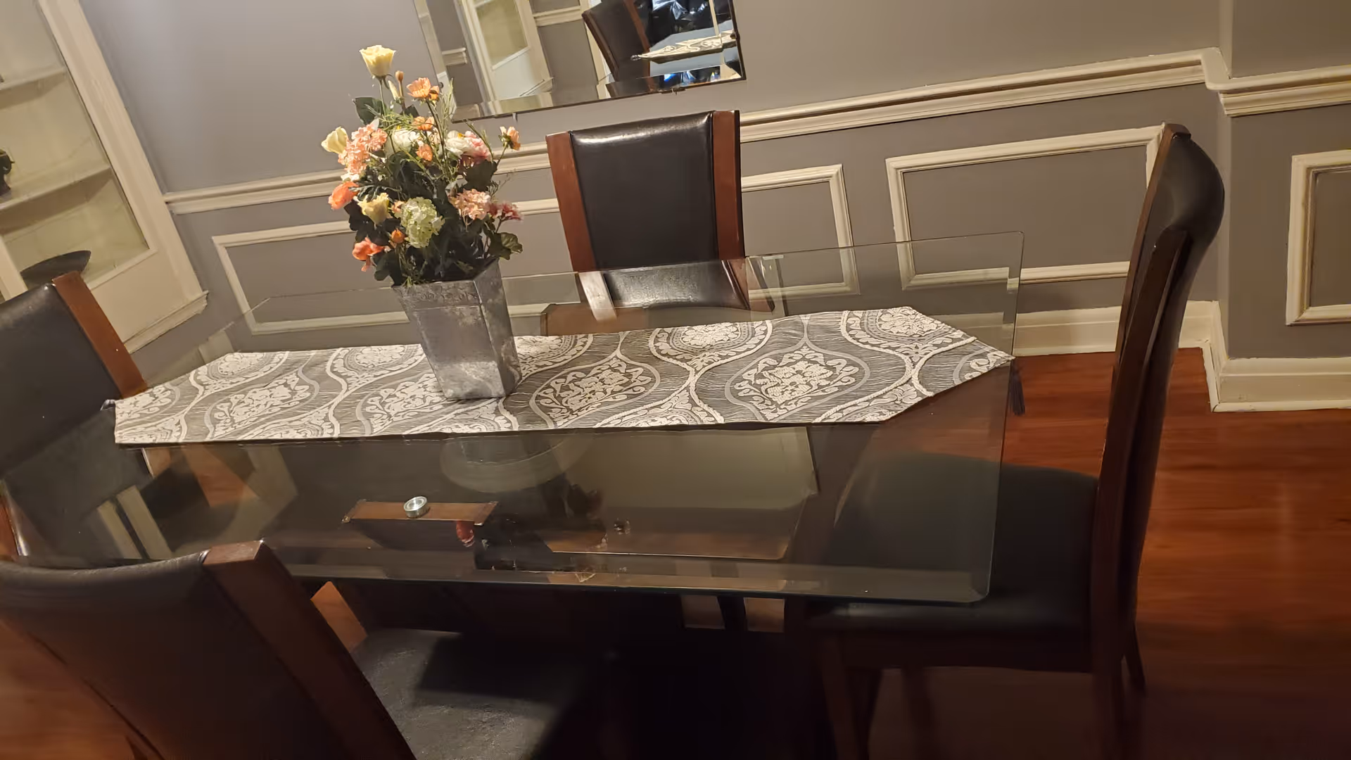 A dining room with a glass-top table and four dark brown chairs with leather upholstery. A decorative table runner with a gray and white pattern is placed on the table, and a metal vase with a bouquet of artificial flowers sits in the center. The room has wooden flooring, gray walls with white trim, and a large mirror on the wall behind the table.