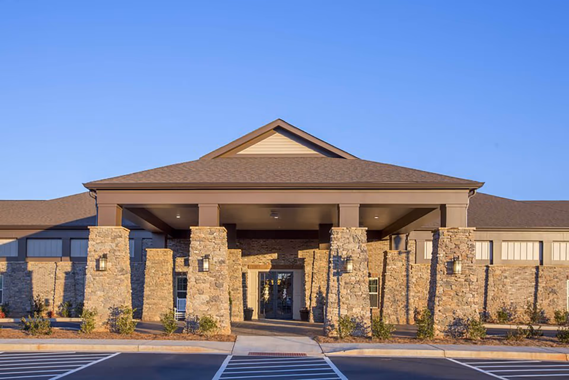 Front exterior view of a senior living facility building with stone pillars supporting a covered entrance, surrounded by small shrubs and a clear blue sky.