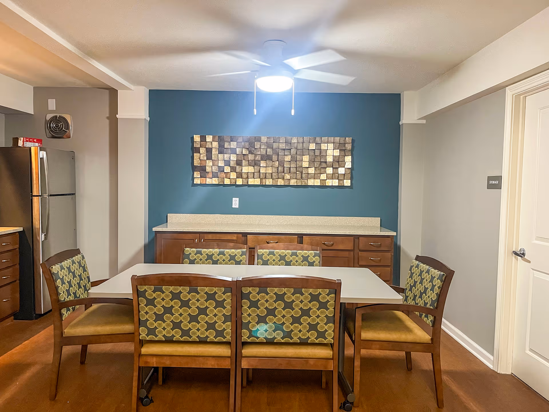 A dining area in a senior living facility with a rectangular table surrounded by six wooden chairs with patterned upholstery. Behind the table is a countertop with wooden cabinets below and a decorative wall art piece above. A ceiling fan with a light is mounted on the ceiling. To the left, a stainless steel refrigerator is visible, and to the right, a door labeled 'STORAGE' is seen.