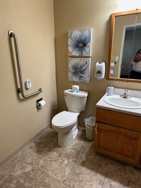 Small accessible bathroom with a toilet, grab bar, wall-mounted soap dispenser, sink with vanity and mirror, floral wall art, and a trash can.