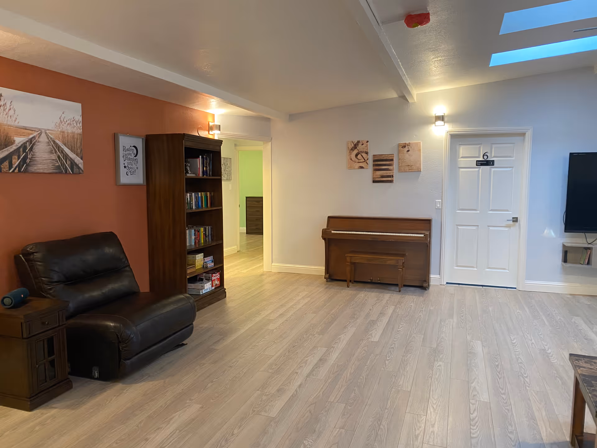 A cozy living room area with light wood flooring, a dark brown leather armchair, a wooden bookshelf filled with books and games, a wooden piano with a matching bench, and a white door labeled with the number 6. The walls are painted in a combination of warm orange and light gray, decorated with framed pictures and wall-mounted lights. A flat-screen TV is partially visible on the right side, and a skylight allows natural light into the room.