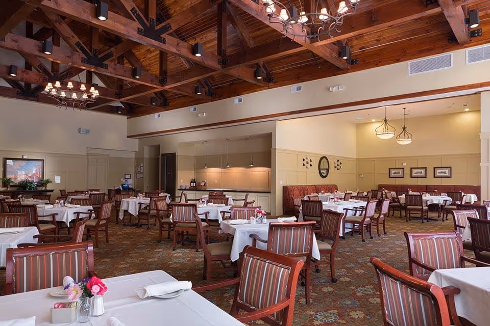 Spacious dining room with multiple white‑clothed tables, wooden chairs, and exposed wooden ceiling beams.