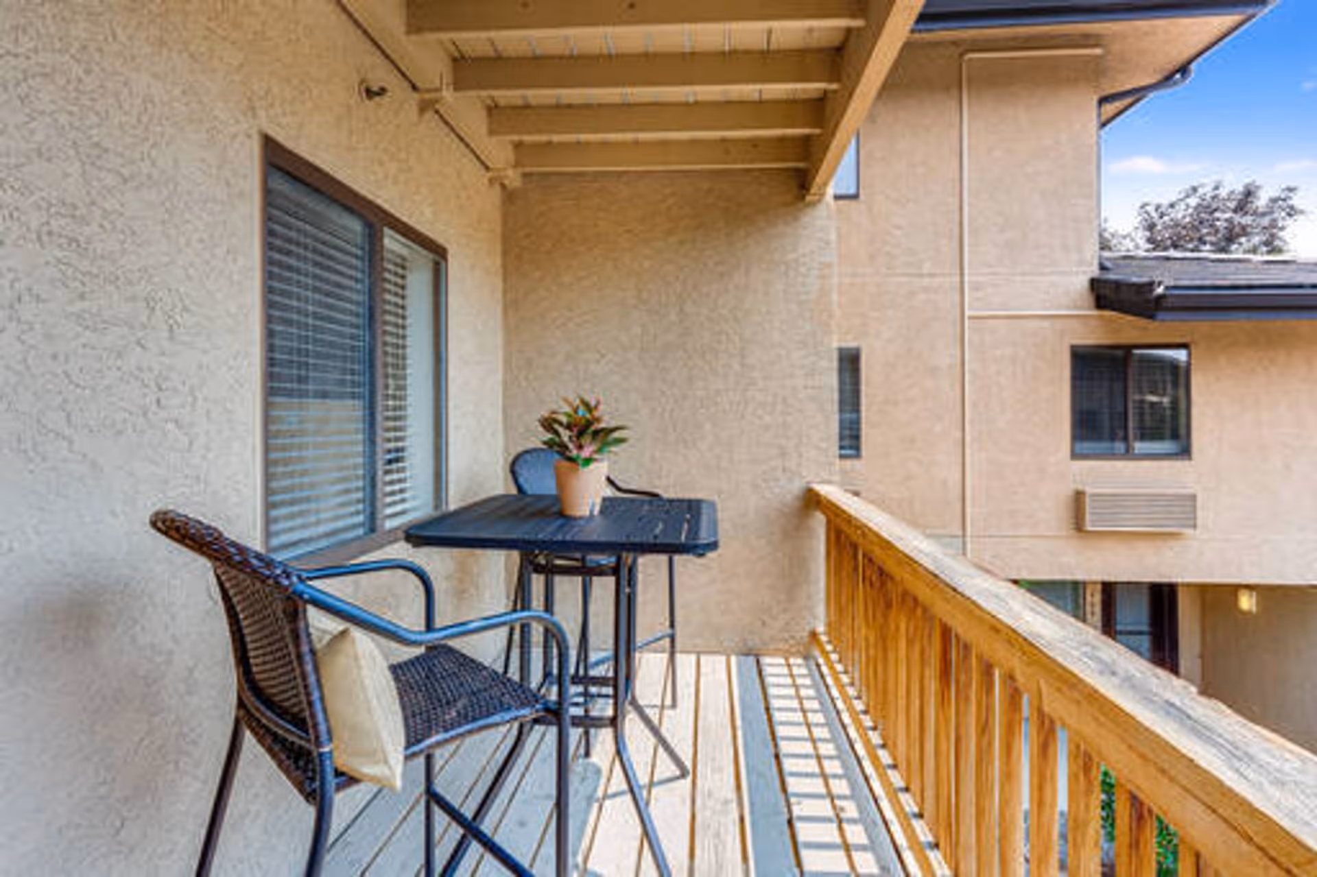 Covered balcony with a small table, two wicker chairs and a potted plant overlooking the neighboring building.