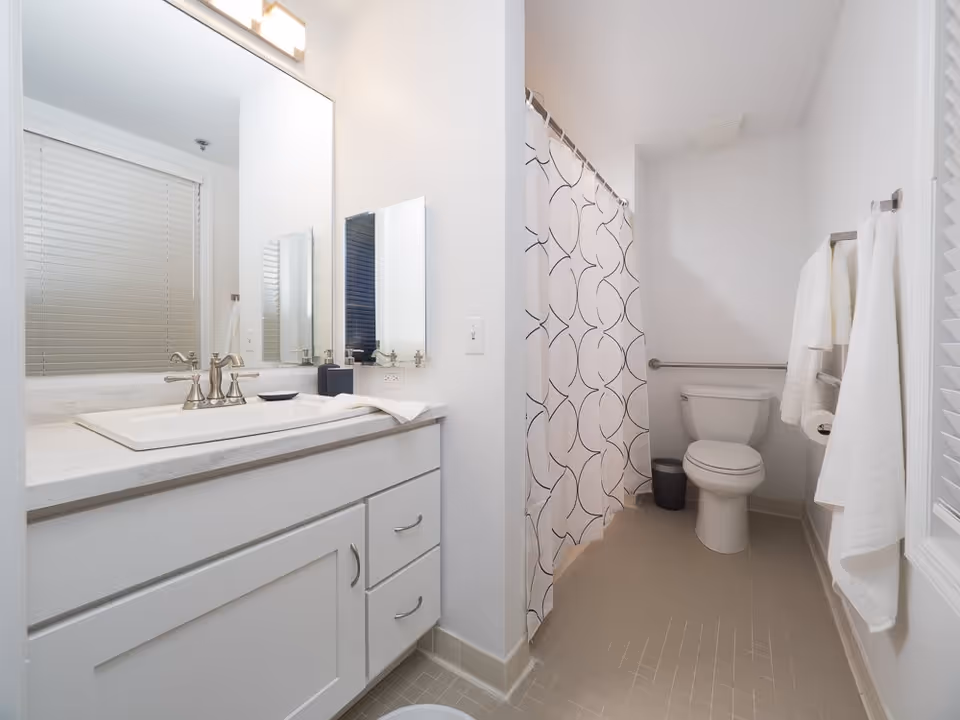 A clean, modern bathroom with a white vanity and sink on the left, a large mirror above the sink, a toilet in the back right corner, and a shower with a white curtain featuring a black circular pattern. White towels hang on a rack on the right wall.