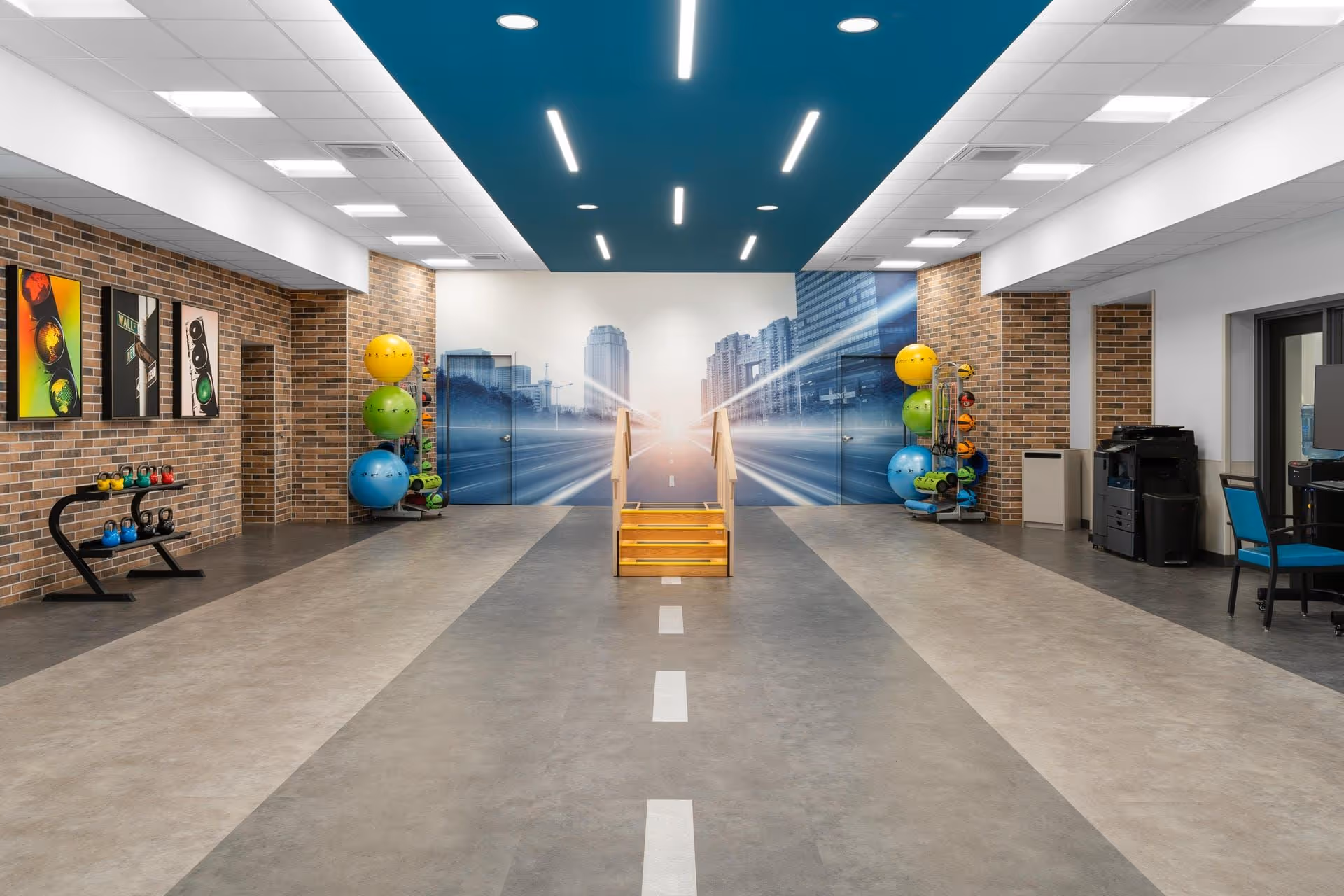 Bright indoor therapy/exercise room with a central walkway and small wooden stairs, exercise balls, kettlebells, brick walls and a cityscape mural.