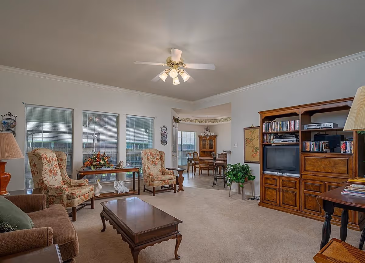 Bright living room with floral upholstered armchairs, a wooden coffee table, an entertainment center with TV and bookshelves, and a dining area visible in the background.