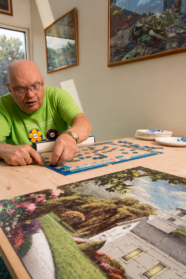 An elderly man wearing glasses and a green shirt is sitting at a wooden table working on assembling a jigsaw puzzle. The puzzle depicts a scenic image of a white building with a garden. The room has light-colored walls with framed pictures hanging, and sunlight is coming through a window.