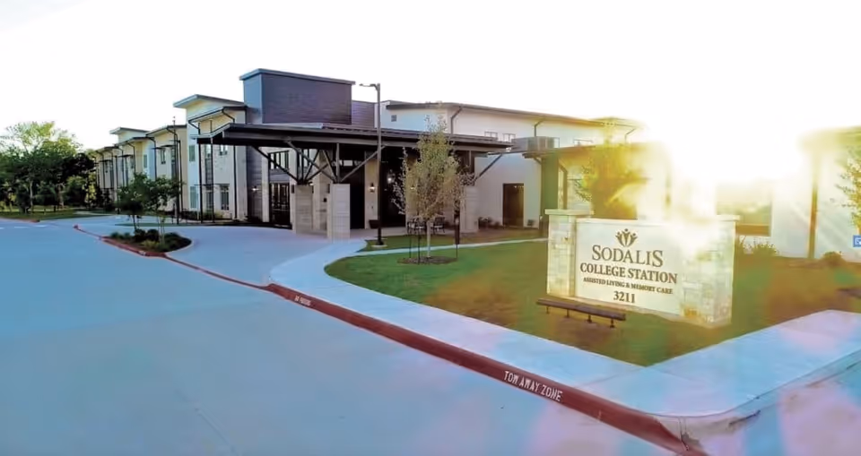 Exterior view of Sodalis College Station assisted living and memory care facility with a large sign in front displaying the facility name and address 3211. The building is modern with multiple windows and a covered entrance. The sun is shining brightly in the background.
