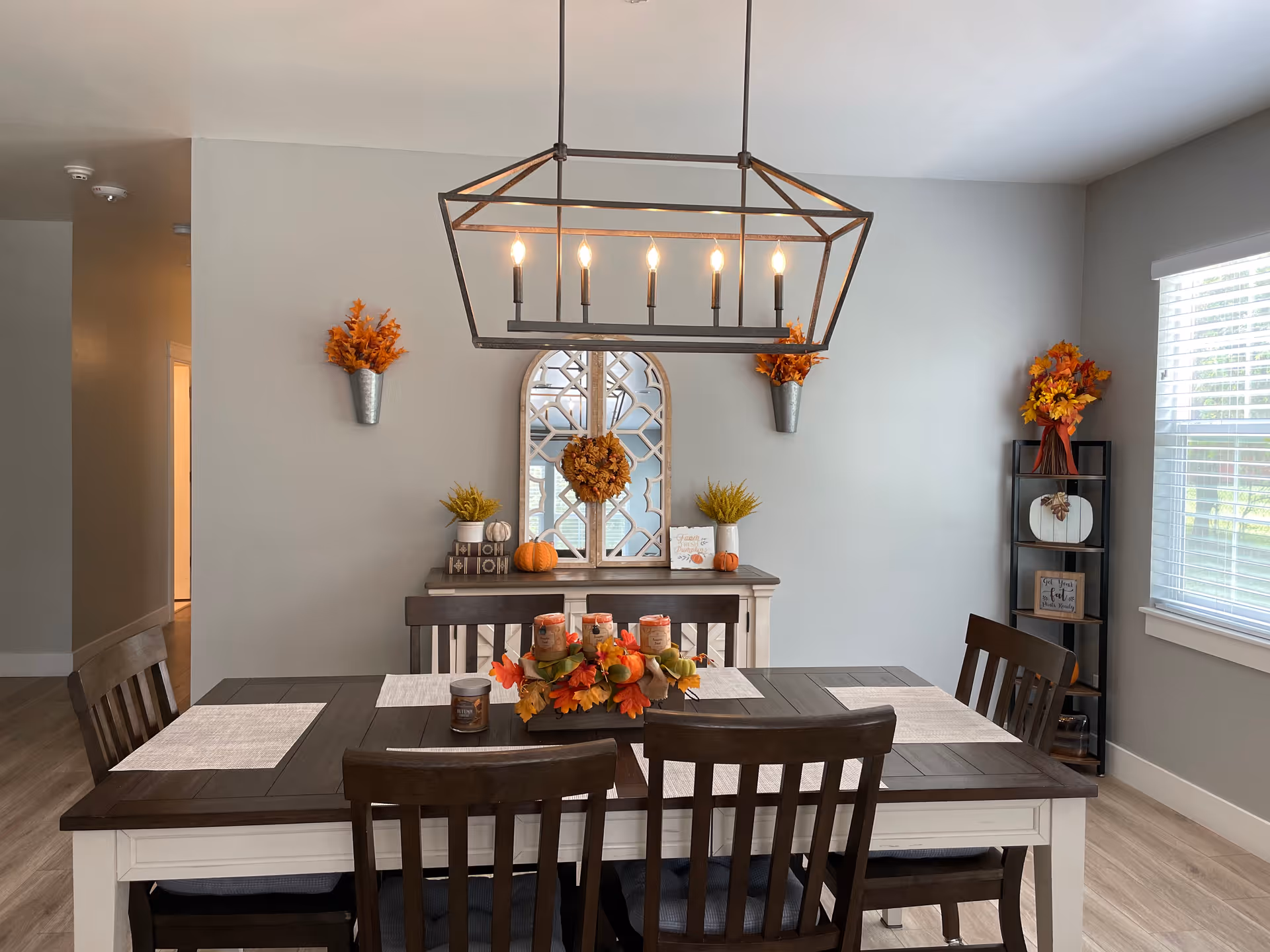 A dining room with a dark wood table and four matching chairs. The table is decorated with a centerpiece featuring autumn leaves and candles. On the wall behind the table is a console table with decorative items including small pumpkins, plants, and a mirror with a wreath. Two wall-mounted metal planters with orange foliage flank the mirror. To the right, a black shelving unit holds more autumn-themed decorations near a window with white blinds. A modern rectangular chandelier with six lit bulbs hangs above the table.