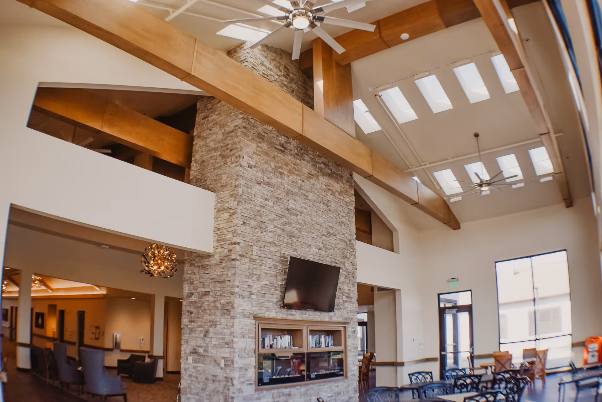 Spacious senior living common room with a tall stone fireplace topped by a wall-mounted TV, high beamed ceiling with skylights, and seating and dining tables.