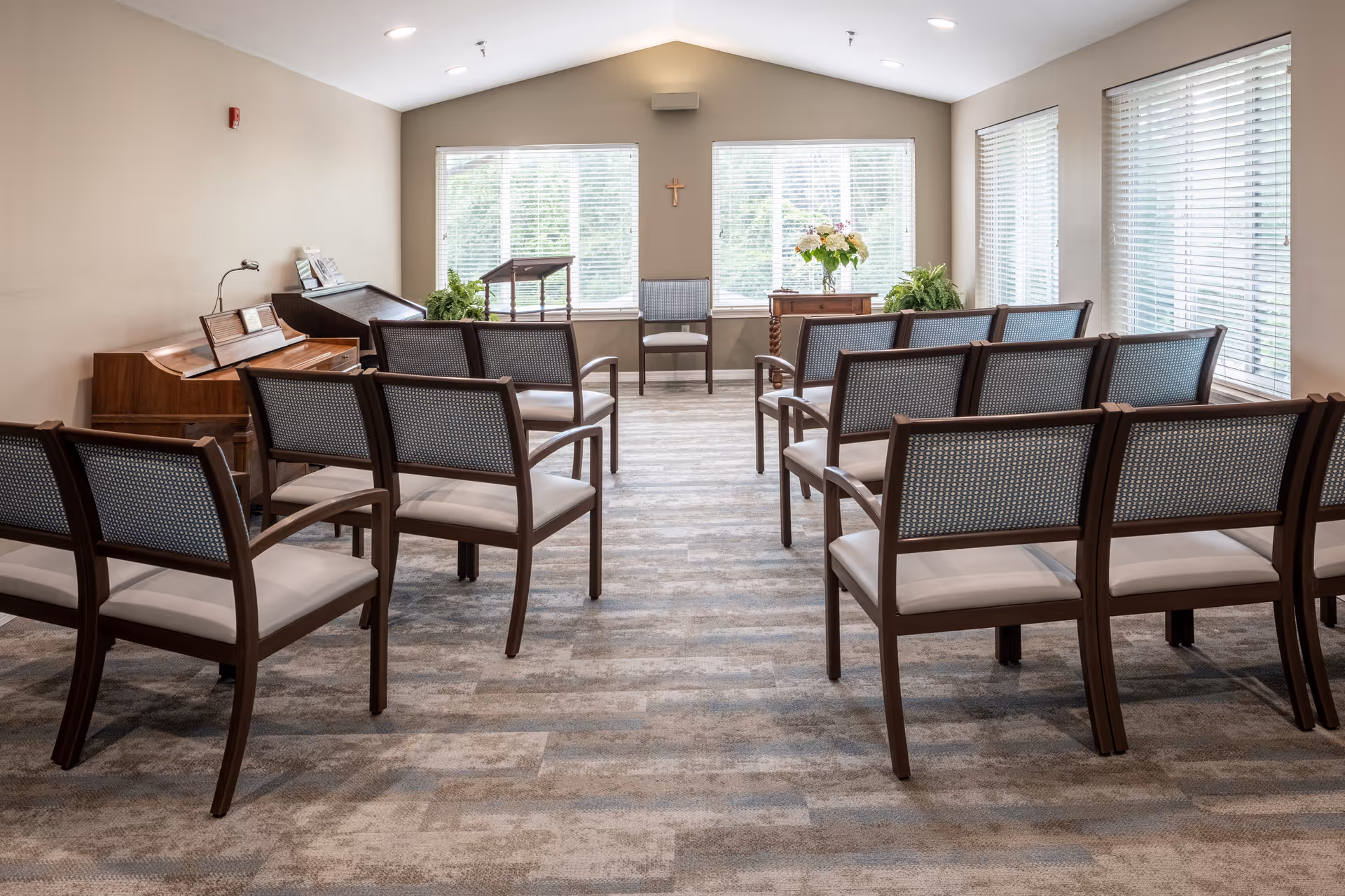 A small chapel or meditation room with rows of wooden chairs with cushioned seats arranged facing a small wooden podium and a single chair at the front. The room has large windows with blinds letting in natural light, a piano on the left side, and plants and flowers near the windows. A small cross is mounted on the wall between the windows.