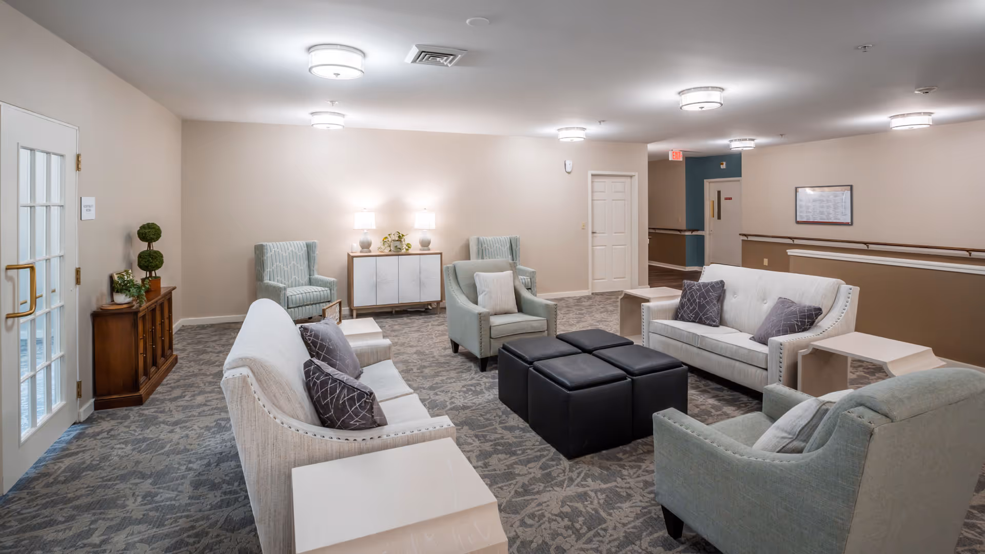 A well-lit senior living facility common area with two white sofas, two light green armchairs, and four black ottomans arranged in the center. The room has beige walls, patterned carpet, and ceiling lights. There is a wooden cabinet with plants on the left side and a white sideboard with lamps and decorations against the far wall.