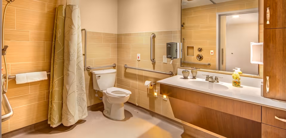 A clean and accessible bathroom with beige tiled walls, a white toilet with grab bars around it, a shower area with a floral patterned curtain, a large mirror above a wooden vanity with a white countertop, soap dispenser, and decorative items.