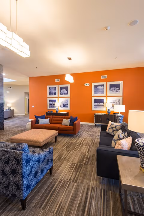 A modern living room area with a bright orange accent wall featuring six framed black and white landscape photographs. The room has a brown sofa with blue and gray pillows, a blue patterned armchair, a dark gray sofa with patterned pillows, and a large rectangular ottoman. There are two table lamps with blue and white bases on dark wood side tables, and two pendant lights hanging from the ceiling. The floor is covered with a striped carpet in neutral tones.