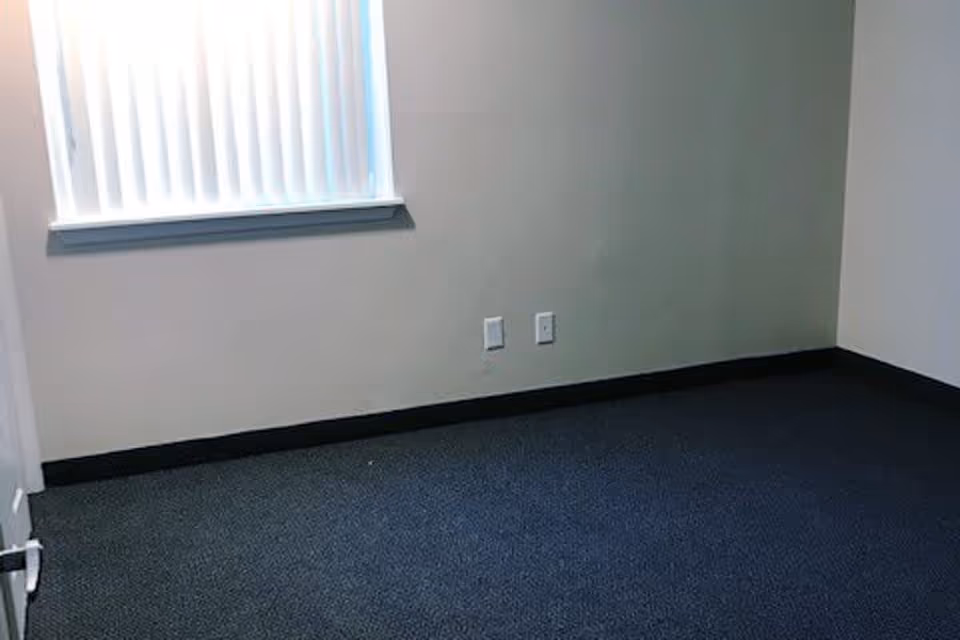Empty room with dark carpet flooring, beige walls, a window with vertical blinds partially open, and two electrical outlets on the wall.