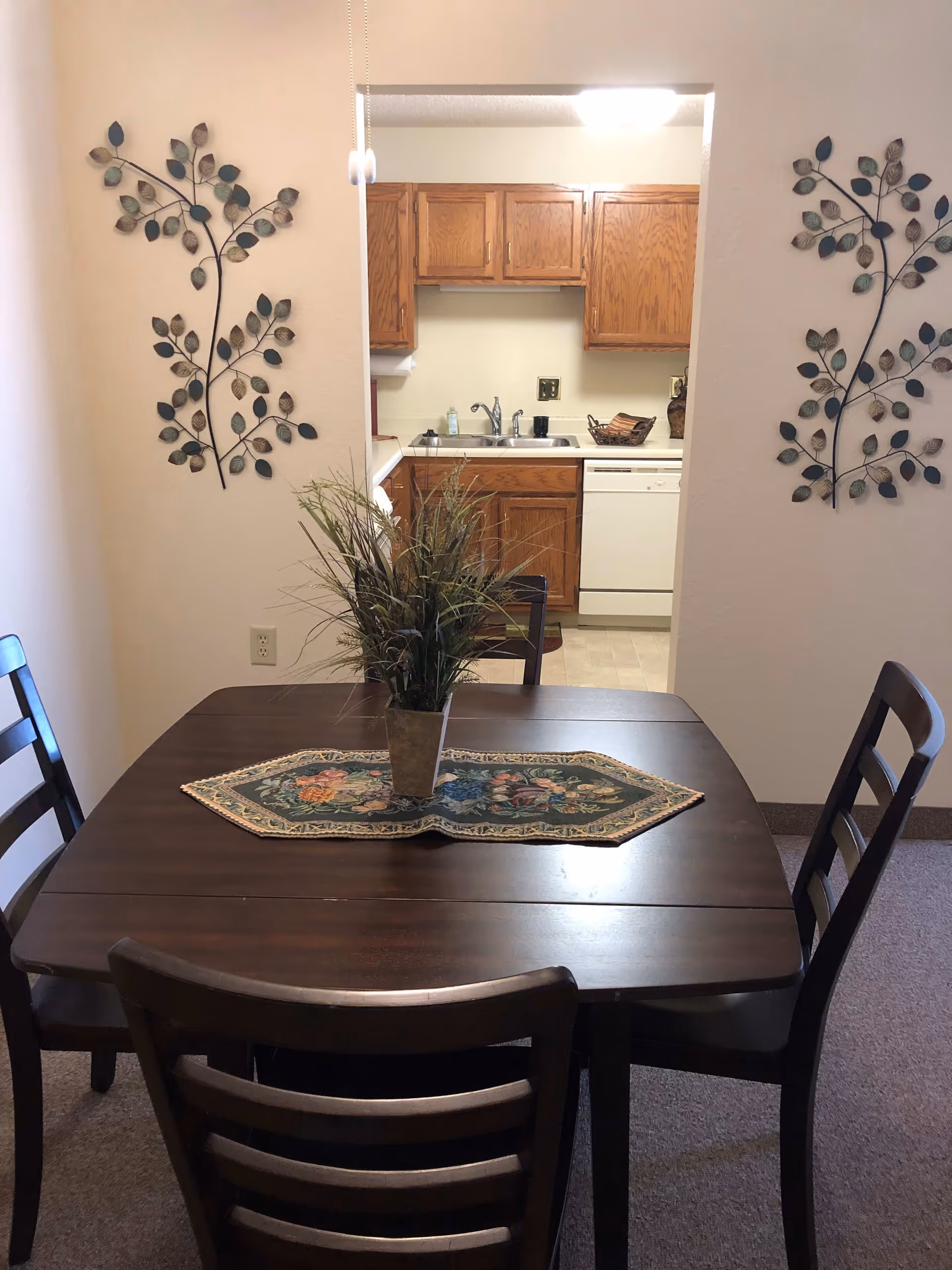 Small dining area with a dark wood table and four chairs, a floral runner and centerpiece, and a view into a kitchen with oak cabinets.