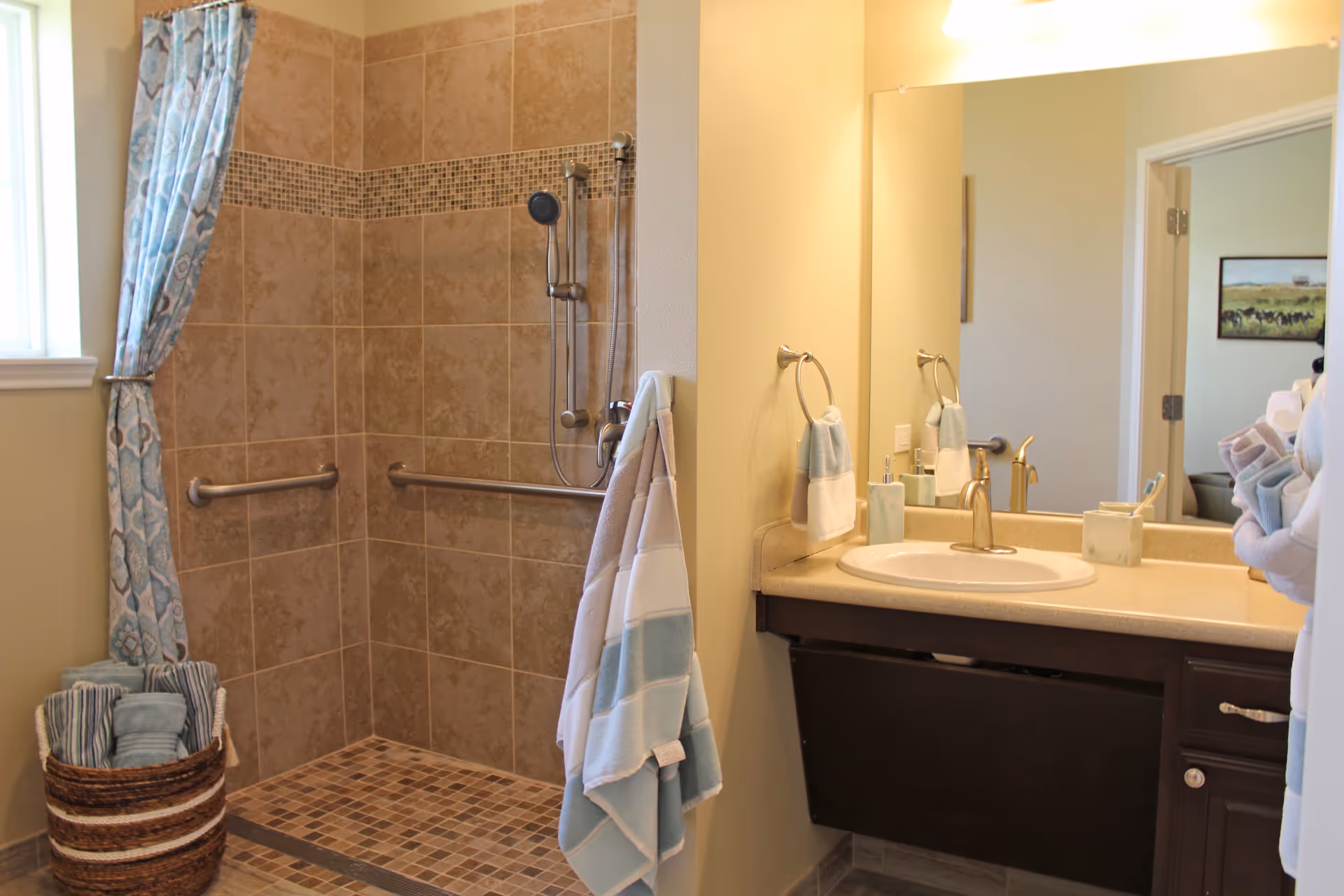 A bathroom with a tiled walk-in shower featuring grab bars and a handheld showerhead. There is a window with a patterned curtain, a basket with rolled towels, and a vanity with a sink, mirror, and neatly arranged towels and toiletries.