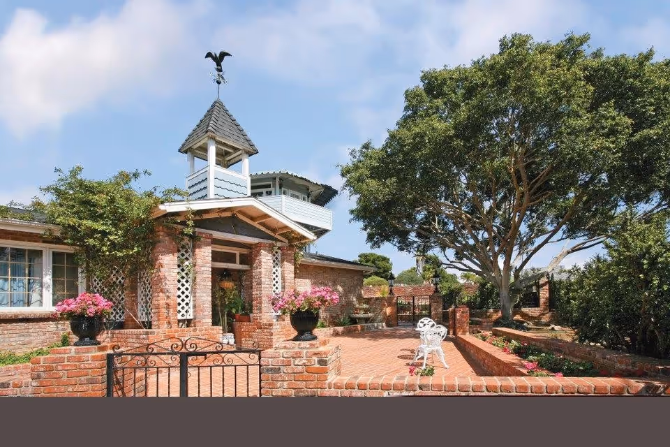 Exterior view of a brick building with a small tower topped by a weather vane. The entrance is framed by brick pillars and lattice panels, with potted pink flowers on either side. There is a brick patio area with a white metal chair and a large tree providing shade. The sky is partly cloudy.