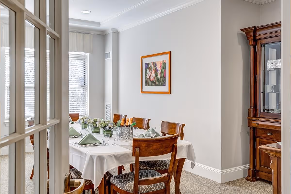 A dining room with a rectangular table covered with a white tablecloth, set with green napkins, glasses, and a centerpiece of flowers and bottles in an ice bucket. The room has wooden chairs with patterned cushions, a wooden china cabinet, and a framed floral painting on the wall. Natural light comes through two windows with blinds.
