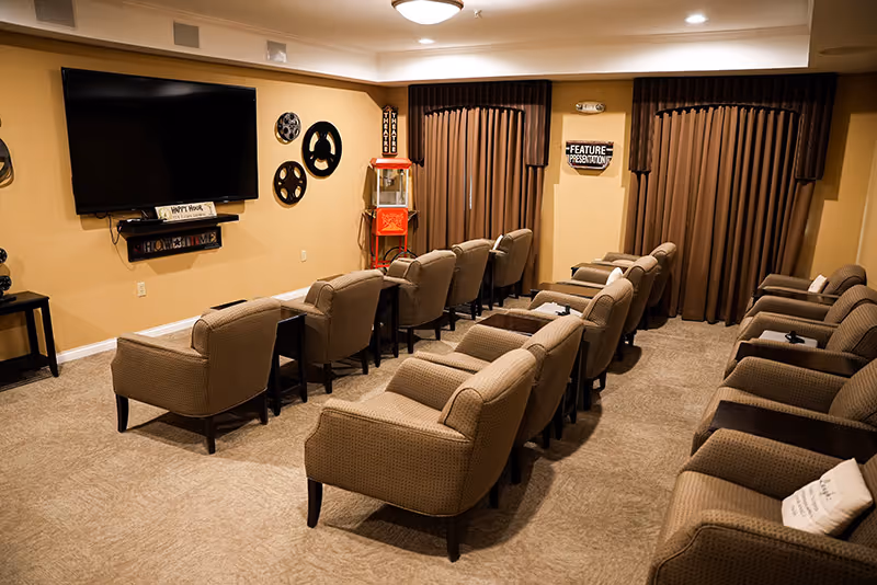 A cozy senior living facility media room with two rows of beige upholstered armchairs facing a large flat-screen TV mounted on a beige wall. The room features movie-themed decor including film reels and a popcorn machine, with brown curtains covering the windows.