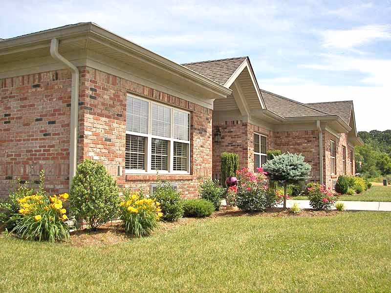 Single-story brick building with white-trimmed windows, a landscaped front lawn with flowering shrubs and a concrete walkway under a partly cloudy sky.