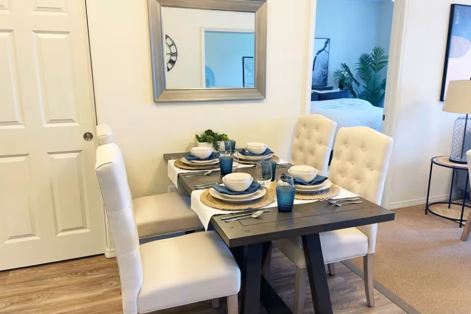 A small dining area with a dark wood table set for four and upholstered tufted chairs next to a mirror and open doorway to a bedroom.