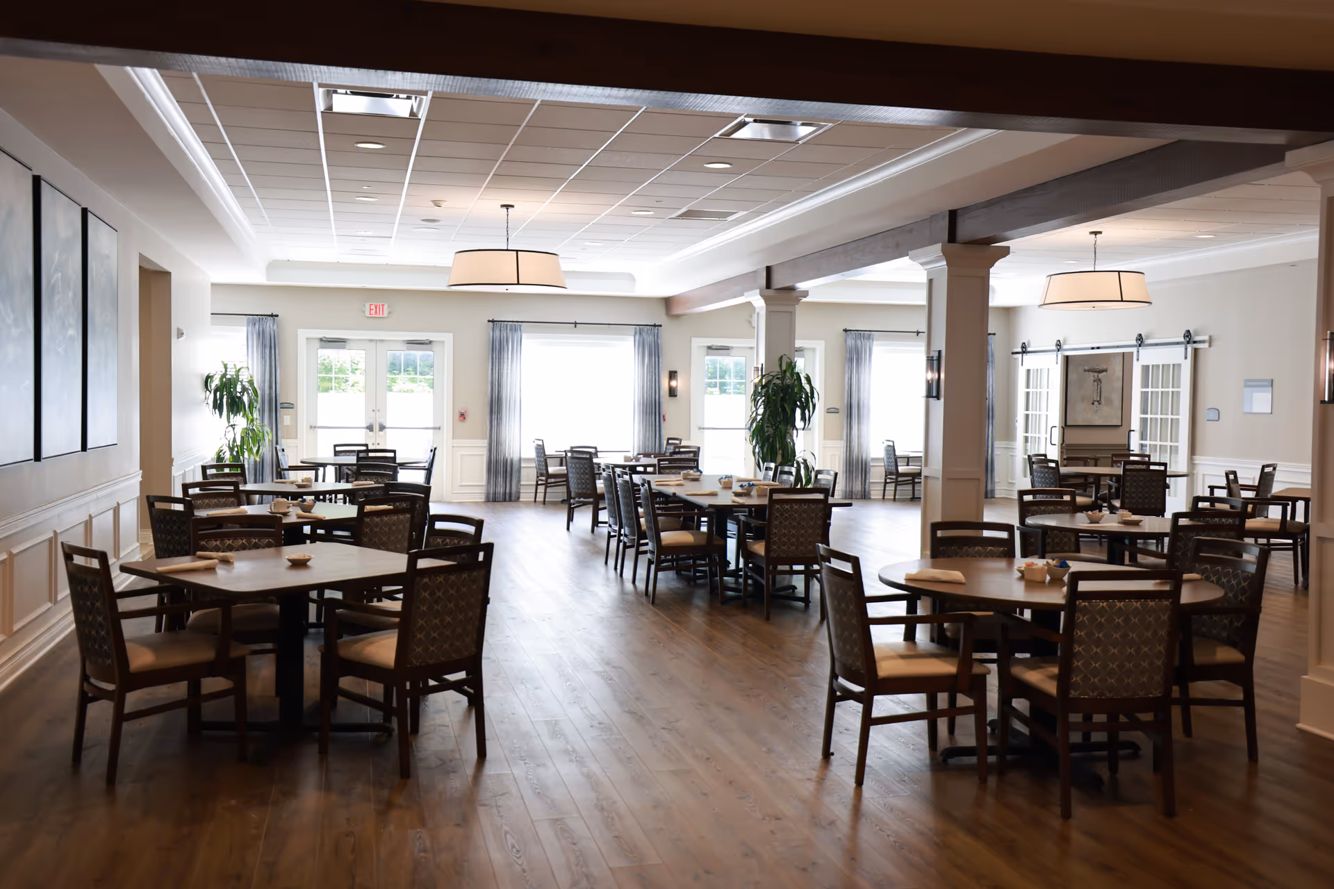 A spacious dining room with multiple round and square wooden tables surrounded by cushioned chairs. The room has large windows with sheer curtains allowing natural light to fill the space. There are two large ceiling light fixtures, wooden flooring, and decorative plants near the windows. The walls are painted light with white trim and some framed artwork.
