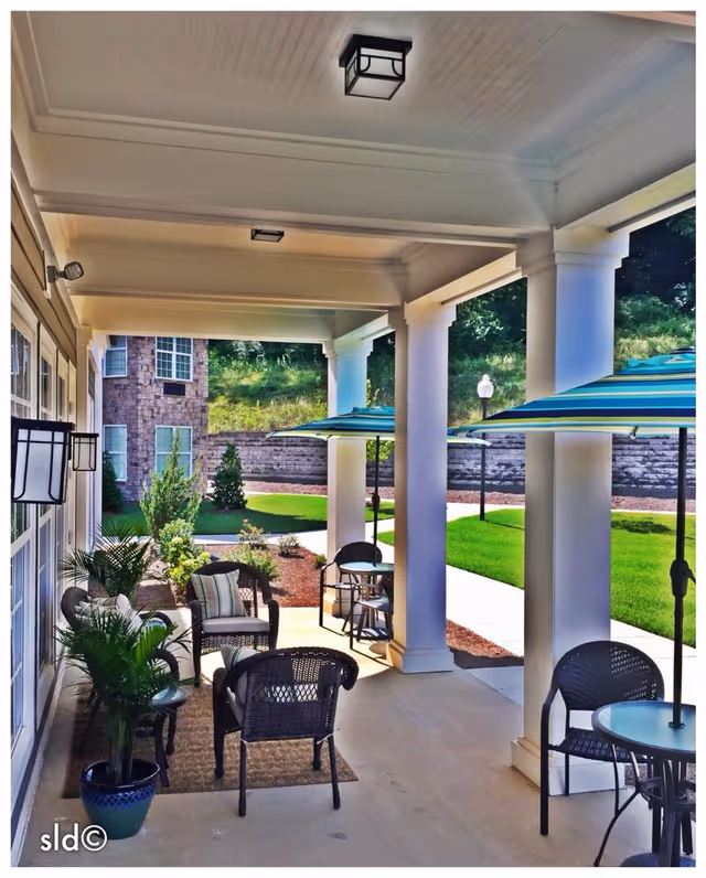 Covered outdoor patio area with white columns, several wicker chairs with cushions, small tables, potted plants, and striped umbrellas providing shade. Green lawn and stone building wall are visible in the background.