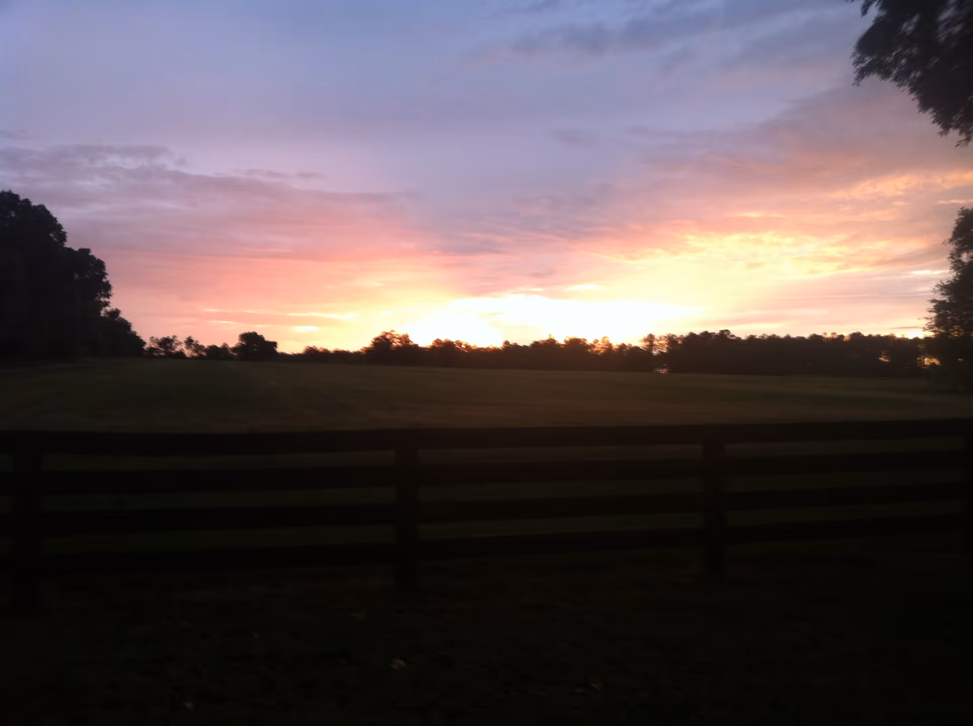 A scenic view of a sunset over an open field with a wooden fence in the foreground and trees silhouetted against the colorful sky.