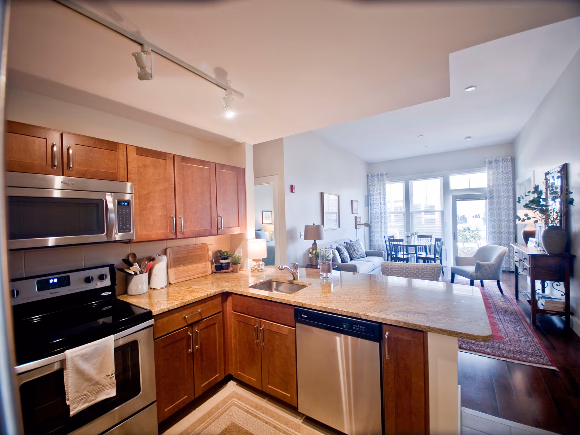 Modern kitchen with wooden cabinets, stainless steel appliances including a microwave, stove, and dishwasher. The kitchen opens up to a living area with a sofa, armchair, dining table, and large windows letting in natural light.