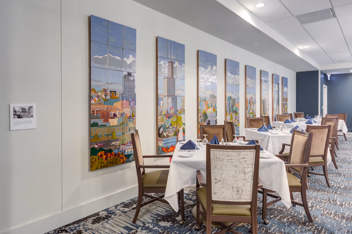 A dining room with several tables covered in white tablecloths, each set with plates, glasses, and blue folded napkins. Wooden chairs with cushioned seats surround the tables. The wall on the left side features a series of colorful tile murals depicting cityscapes. The room has a patterned carpet and a ceiling with recessed lighting.